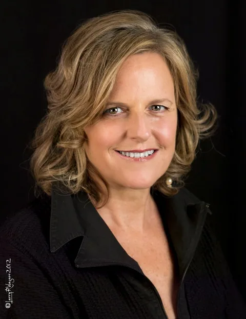 Woman with shoulder-length wavy blonde hair wearing a black collared shirt, smiling at camera against a black background