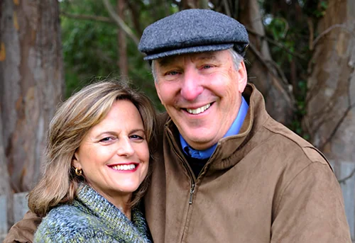 Two smiling adults posing together outdoors in front of wooden posts and greenery