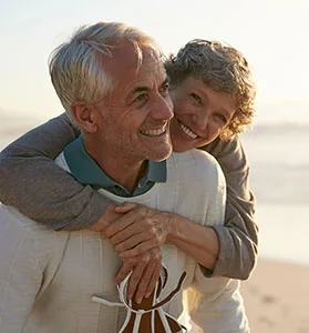 Older man giving piggyback ride to young child on beach at sunset