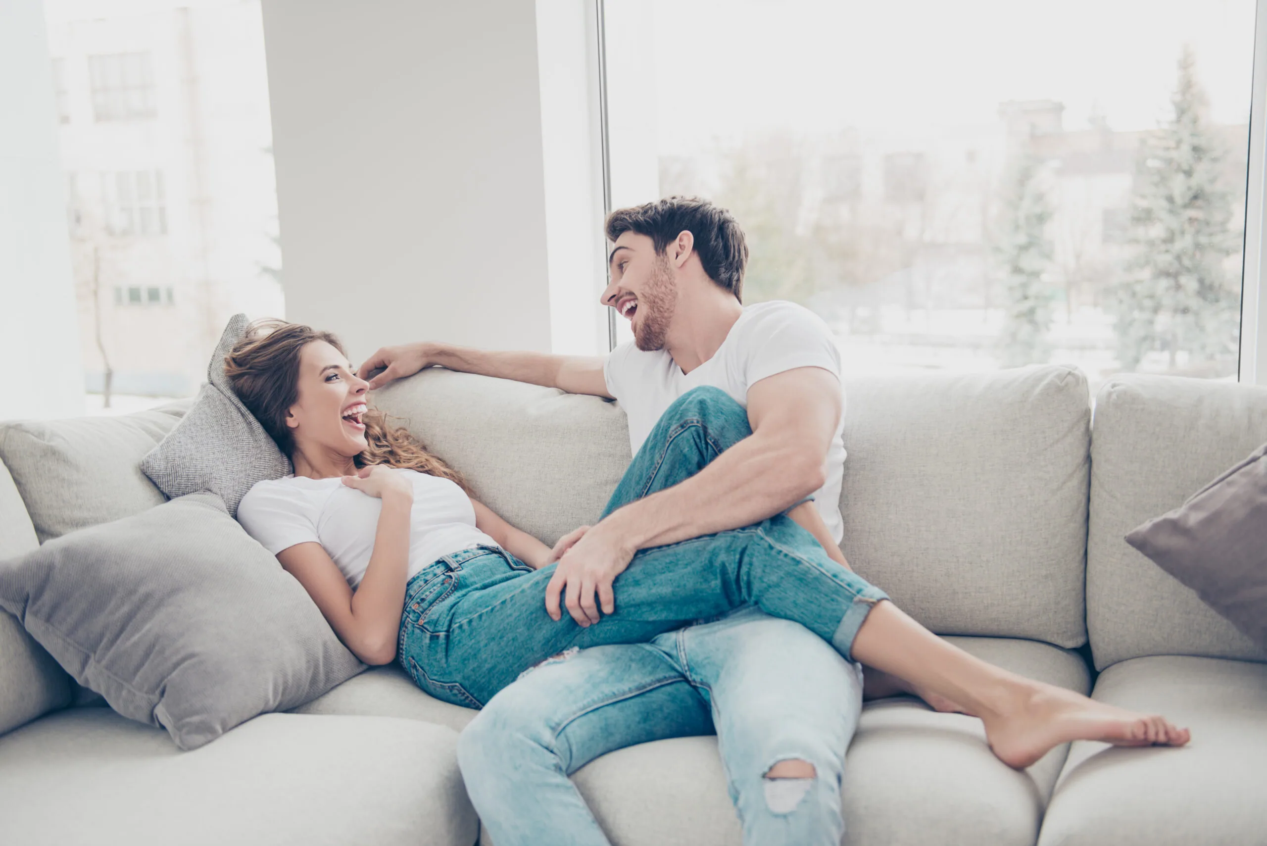 Young couple sitting on light gray couch playfully interacting with city skyline visible through large windows behind them