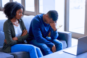 A frustrated husband sits on couch with his head hanging down beside his wife who always seems angry.